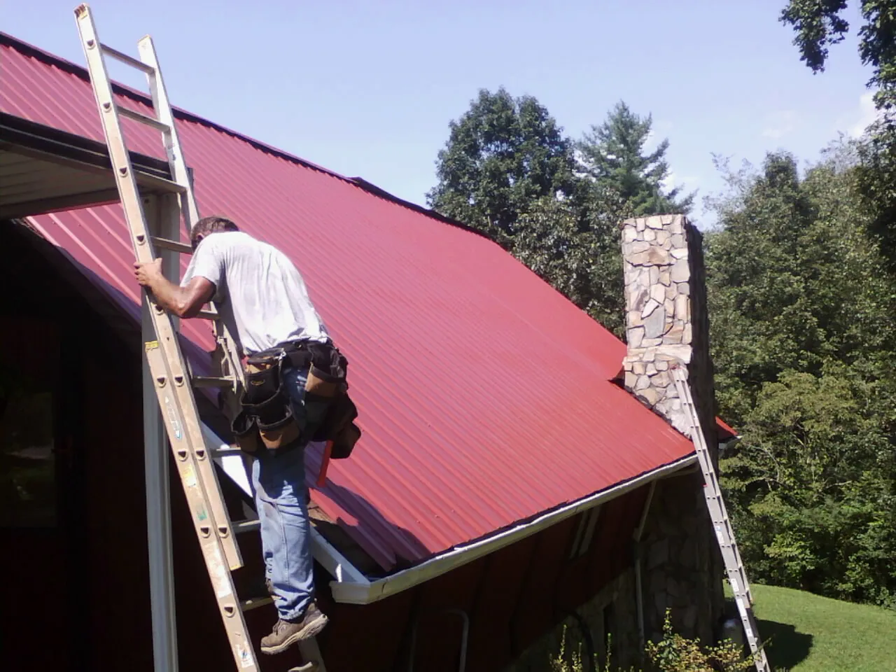 Roofer on a ladder beside a red metal roof and stone chimney — Soffit & Fascia Repair by Roofing Brothers in Bartlesville
